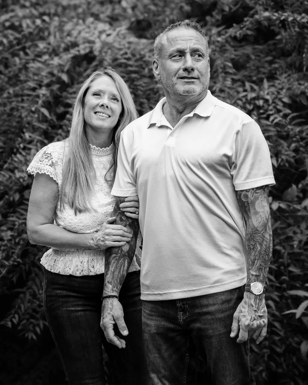 : Black and white photo of a woman with long hair smiling and holding the arm of a tattooed man in a short-sleeve polo shirt and jeans, standing against a leafy background.