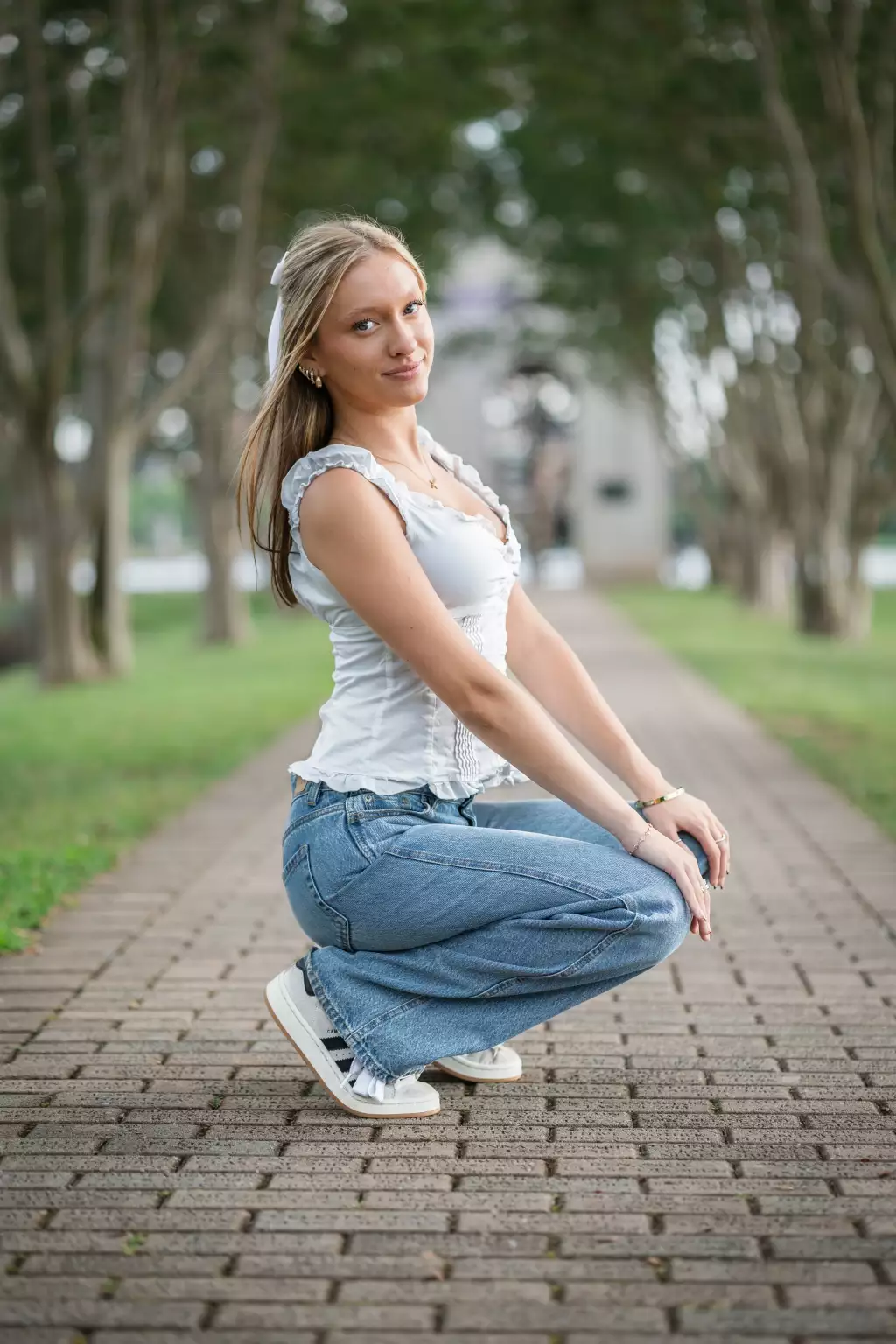 : Young woman with long blonde hair tied with a white ribbon, wearing a white sleeveless ruffled top, blue jeans, and white sneakers, squatting on a brick pathway lined with trees in an outdoor park setting.
