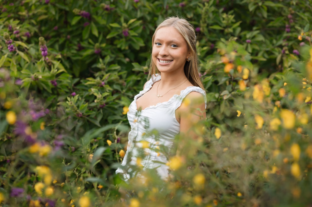 Marlee's senior photos taken at Furman University, Greenville, SC: Young woman with long blonde hair smiling, wearing a white sleeveless top, standing outdoors surrounded by greenery and purple and yellow flowers.