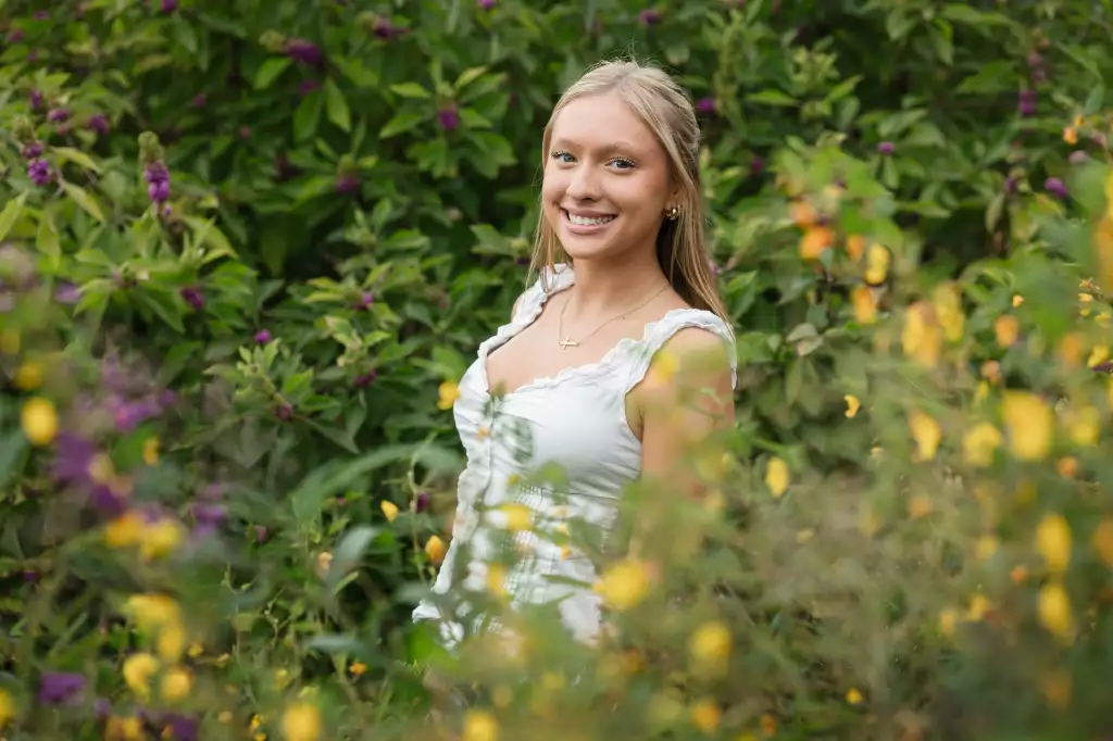 : Young woman with long blonde hair smiling, wearing a white sleeveless top, standing outdoors surrounded by greenery and purple and yellow flowers.