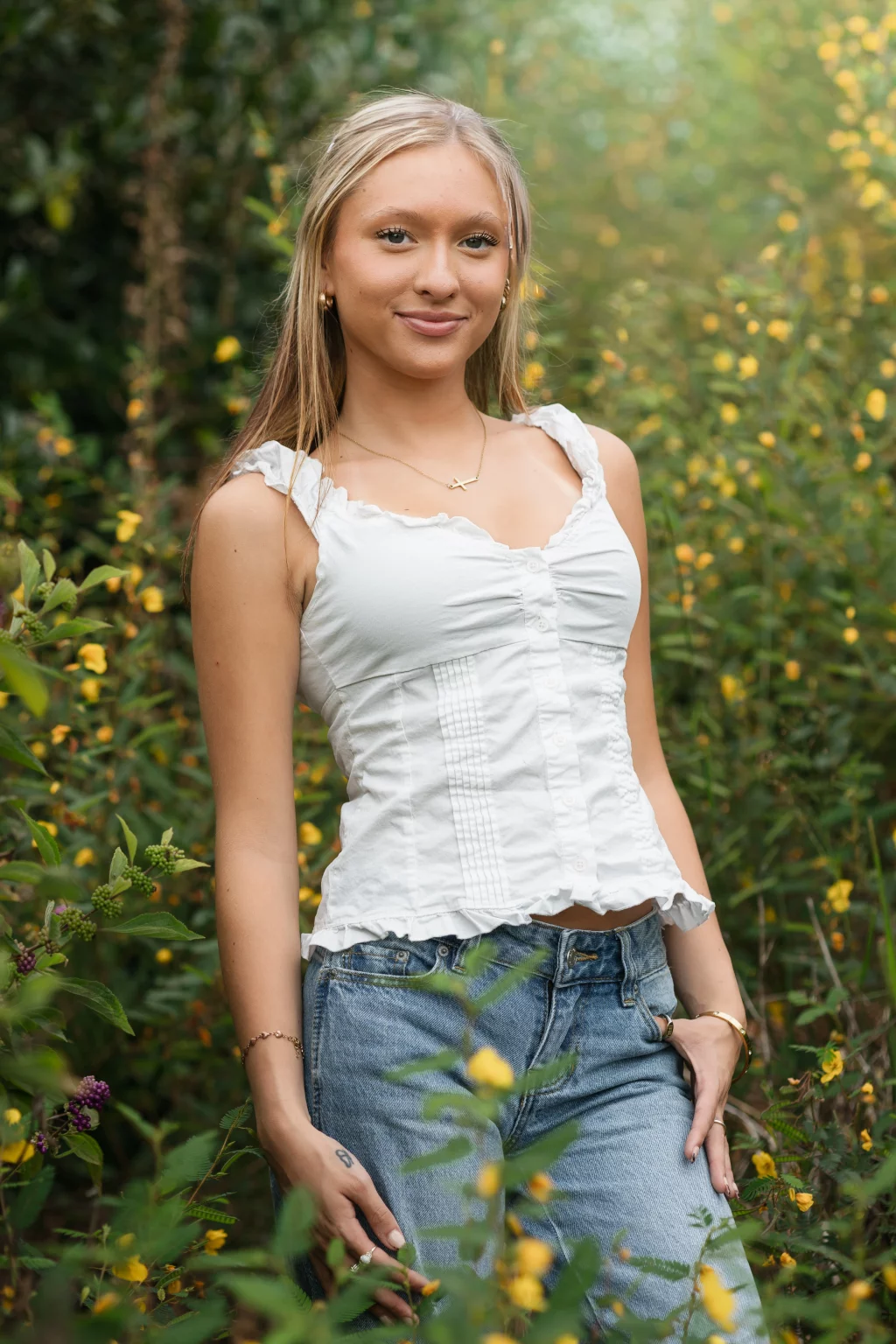 Marlee's senior photos taken at Furman University, Greenville, SC: Young woman with long blonde hair wearing a white sleeveless top and blue jeans standing outdoors among green plants with yellow flowers, smiling gently at the camera.