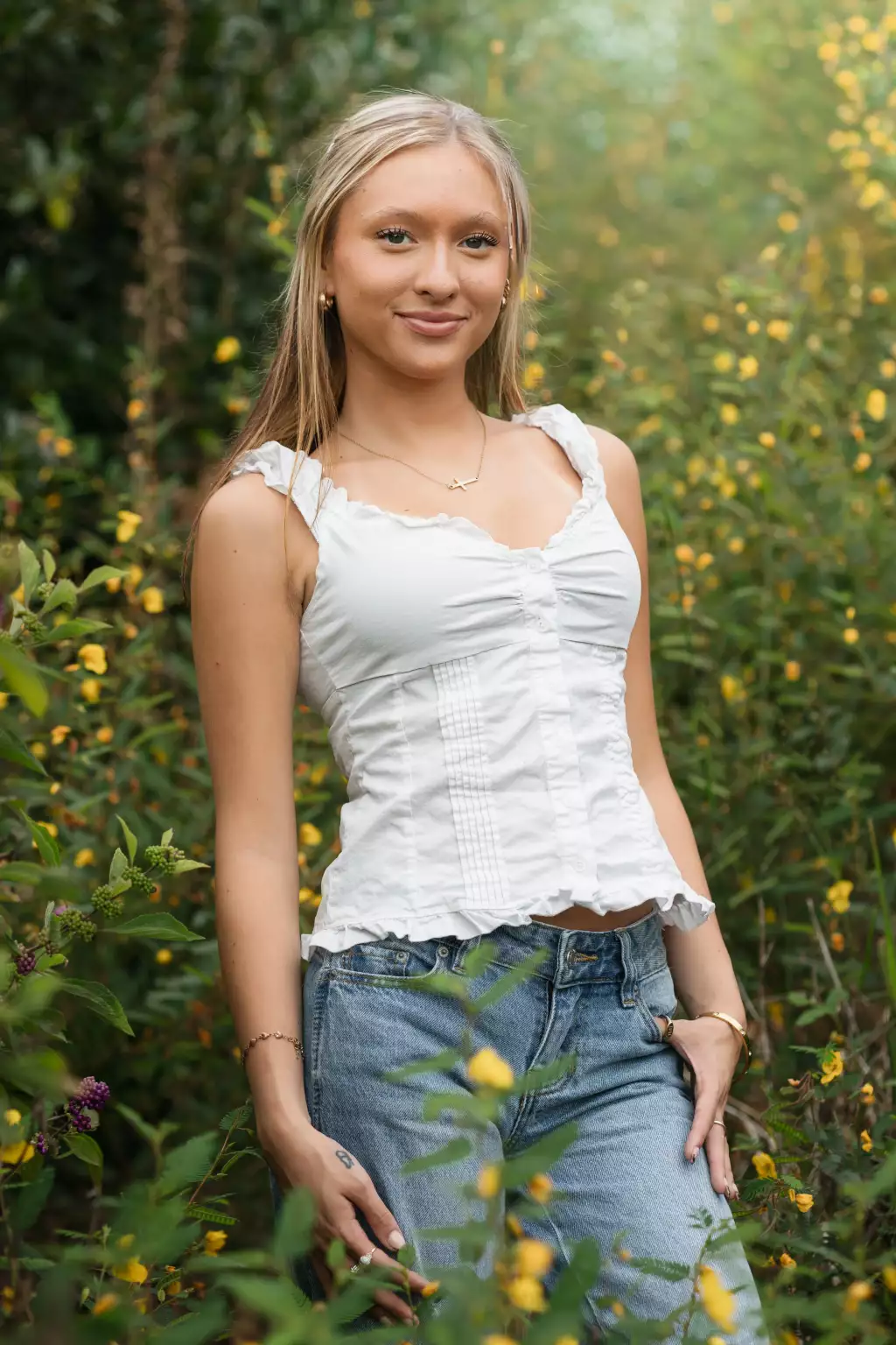 : Young woman with long blonde hair wearing a white sleeveless top and blue jeans standing outdoors among green plants with yellow flowers, smiling gently at the camera.