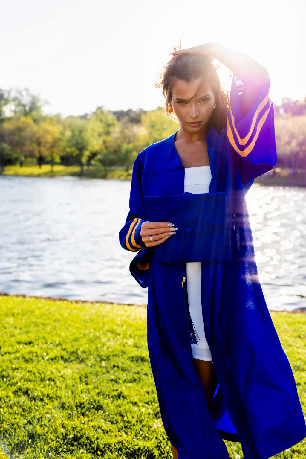 Michelle's senior photos taken at Furman University / Rose garden: Young woman in a blue graduation gown holding a graduation cap, standing outdoors near a body of water with trees in the background, sunlight creating lens flare and backlighting her face.