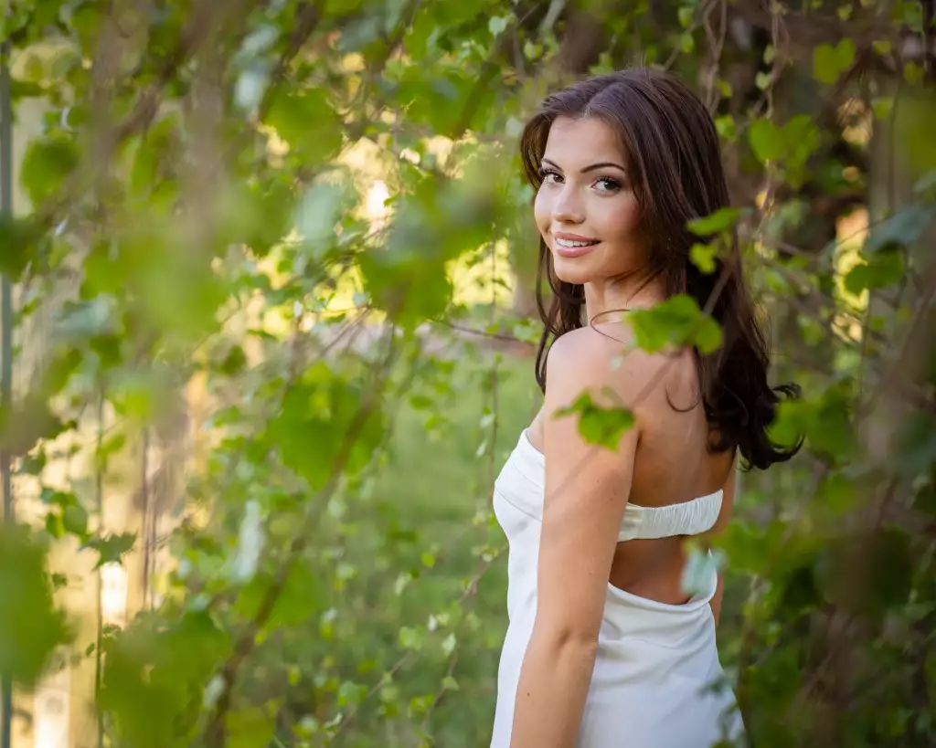 : Smiling woman with long brown hair wearing a white strapless dress, standing outdoors surrounded by green leafy foliage, looking over her shoulder at the camera.
