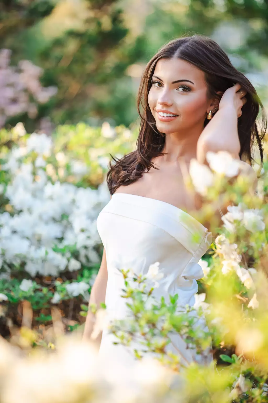 : Young woman with long dark hair wearing a strapless white dress, standing among blooming white flowers and greenery, smiling softly, with one hand touching her hair.