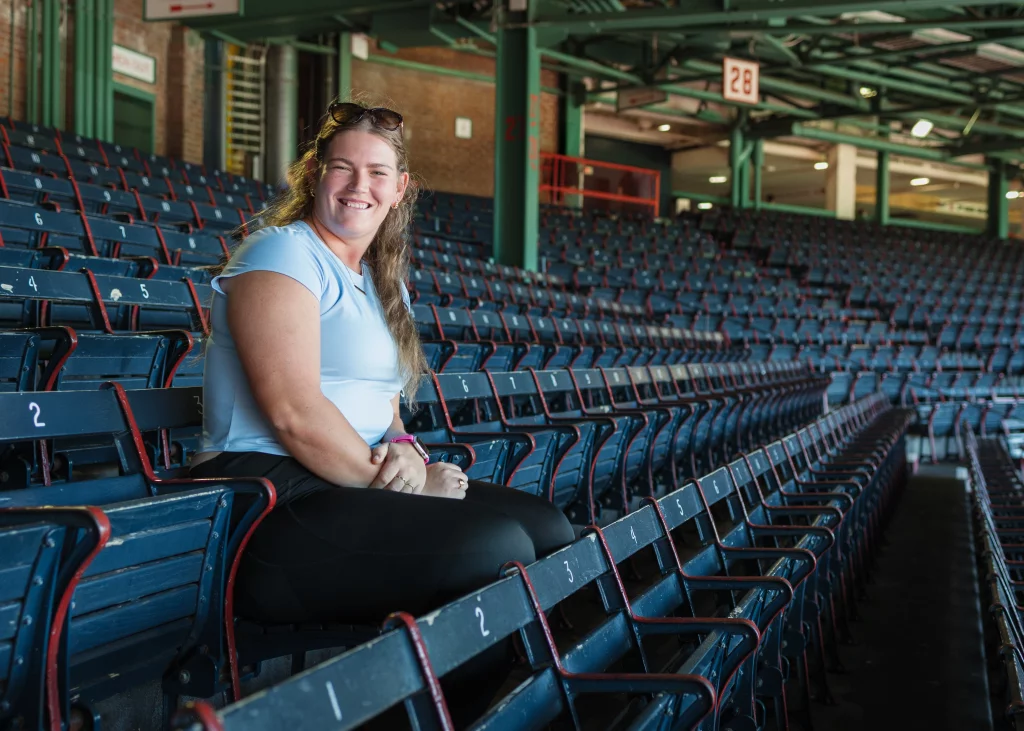 : A woman with long hair and a light blue shirt sits and smiles in an empty section of blue stadium seats.
