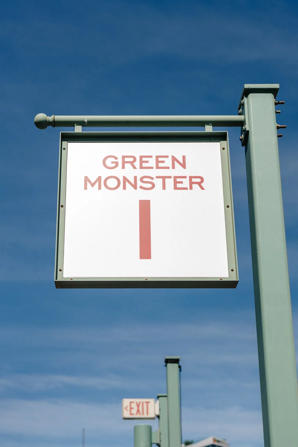 : A sign reading "GREEN MONSTER 1" in red letters, mounted on a green post against a blue sky. An "EXIT" sign is visible in the background.