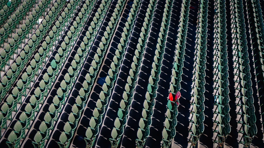 : Rows of empty green stadium seats, with one seat covered by a red cloth standing out among the uniform pattern.
