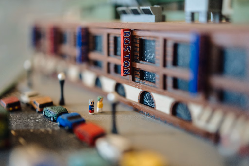 : Close-up of a miniature city scene featuring a model building with a red vertical sign reading "BOSTON," small model cars parked along the street, and two tiny figurines standing on the sidewalk.