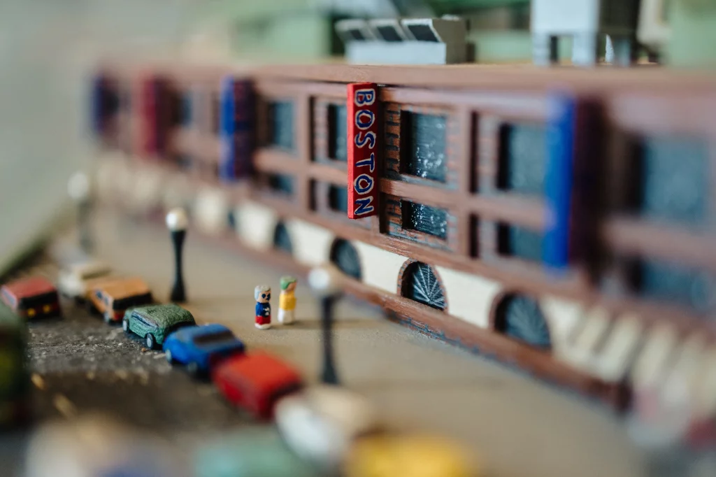 : Close-up of a miniature city scene featuring a model building with a red vertical sign reading "BOSTON," small model cars parked along the street, and two tiny figurines standing on the sidewalk.