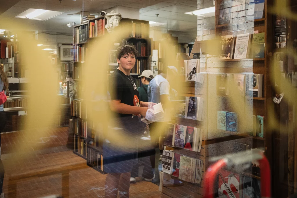 : A young person holding a book stands inside a bookstore, seen through a glass window with large yellow letters. Bookshelves and other people browsing are visible in the background.