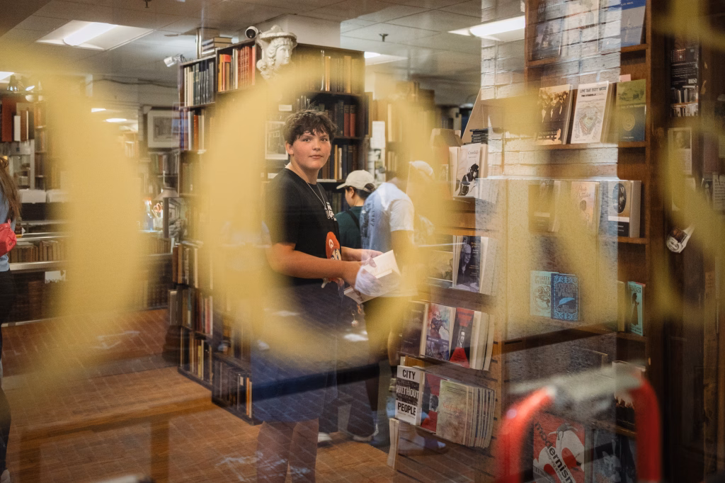 : A young person holding a book stands inside a bookstore, seen through a glass window with large yellow letters. Bookshelves and other people browsing are visible in the background.
