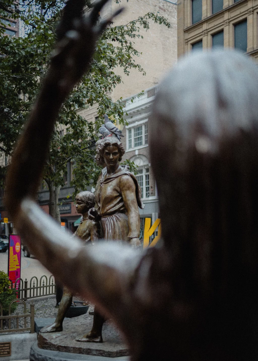 : Bronze statues of a woman and child, with pigeons perched on the woman's head, seen through the raised arm of another statue in the foreground; set in an urban outdoor plaza.