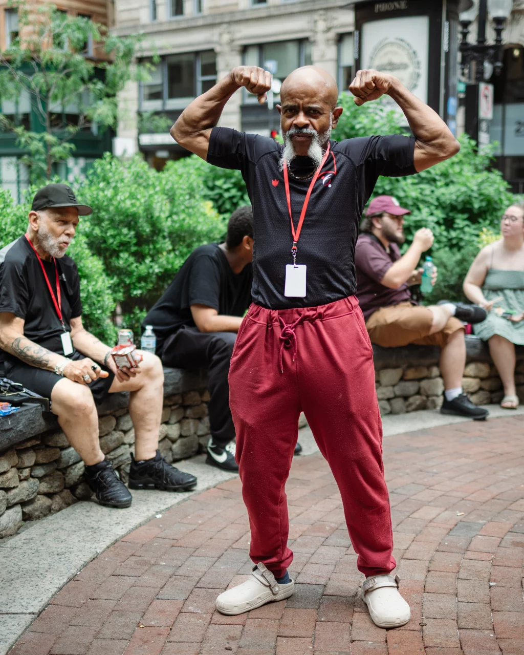 : An older man in a black shirt and red pants stands on a brick walkway, flexing his arms and smiling. Several people sit on a stone wall behind him in a park setting.