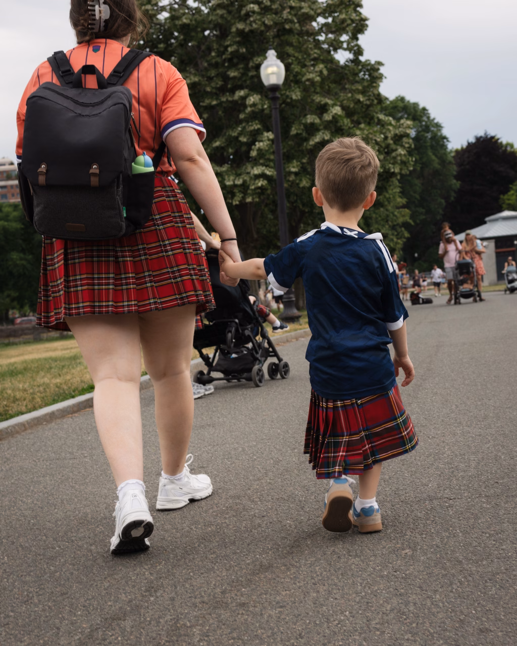 : An adult and a child, both wearing red plaid kilts, walk hand-in-hand down a paved path in a park. The adult carries a black backpack, and the child wears a navy shirt. Other people and strollers are visible in the background.