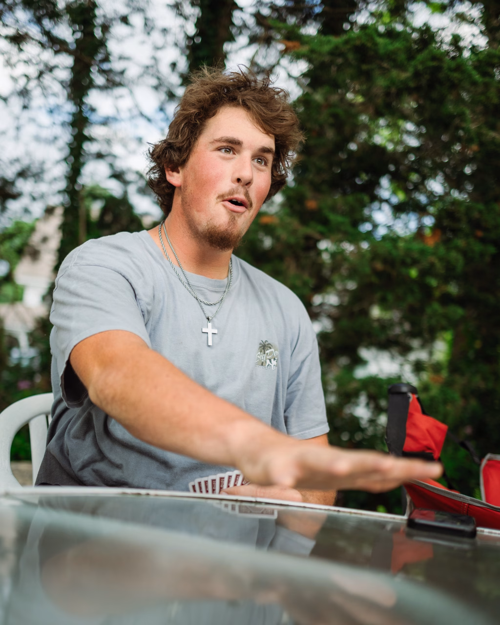 : Young man with light brown hair and a goatee, wearing a gray t-shirt and a cross necklace, sitting at an outdoor table with playing cards in hand, gesturing animatedly with a surprised facial expression. Trees are visible in the background.