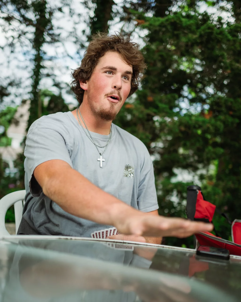 : Young man with light brown hair and a goatee, wearing a gray t-shirt and a cross necklace, sitting at an outdoor table with playing cards in hand, gesturing animatedly with a surprised facial expression. Trees are visible in the background.