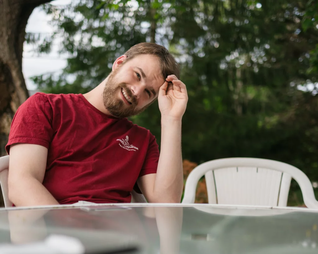 : Man with a beard in a red t-shirt sitting outdoors at a glass table, tilting his head and smiling, with green trees in the background.