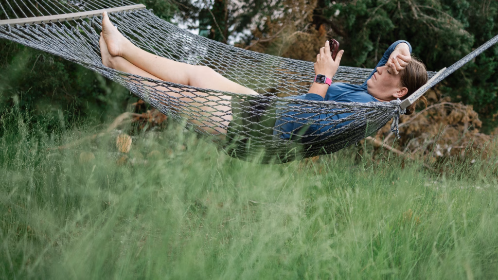 : Person lying in a hammock outdoors, holding a phone with one hand and resting the other hand on their forehead, surrounded by tall green grass and trees.