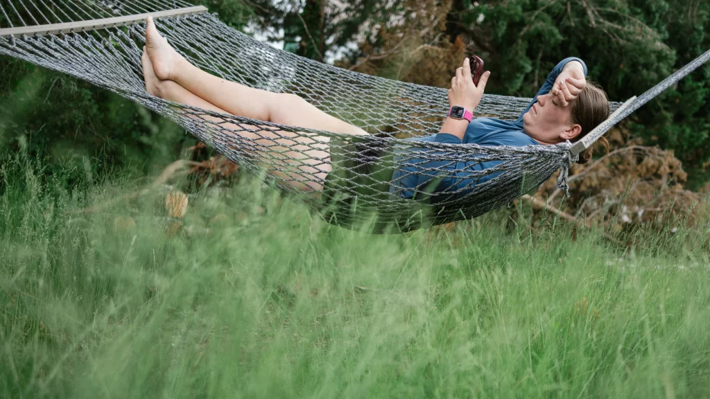 : Person lying in a hammock outdoors, holding a phone with one hand and resting the other hand on their forehead, surrounded by tall green grass and trees.