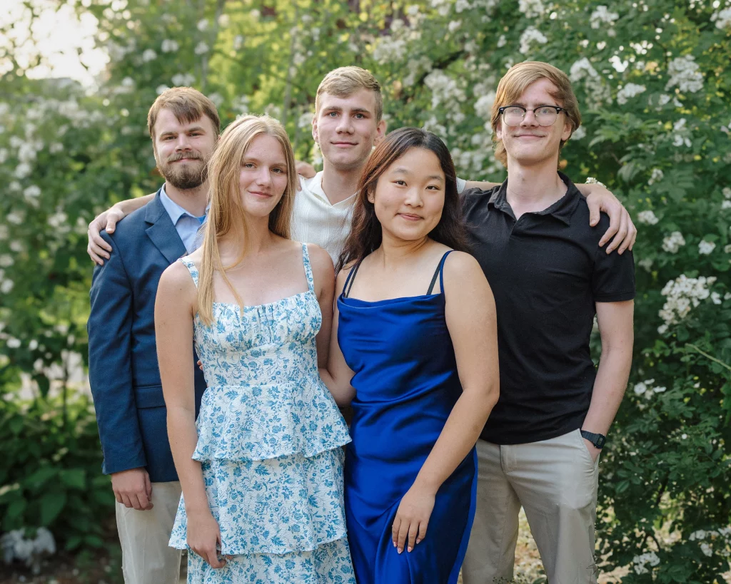 : Five young adults pose together outdoors in front of leafy green bushes with white flowers; two women stand in front wearing dresses, and three men stand behind them, dressed in a mix of casual and semi-formal clothing.