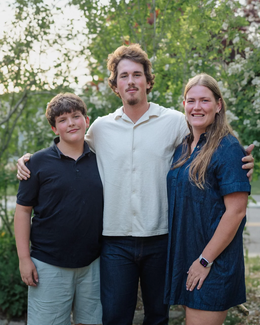 : Three people standing outdoors in front of green foliage, smiling at the camera. The person in the middle has an arm around the other two.