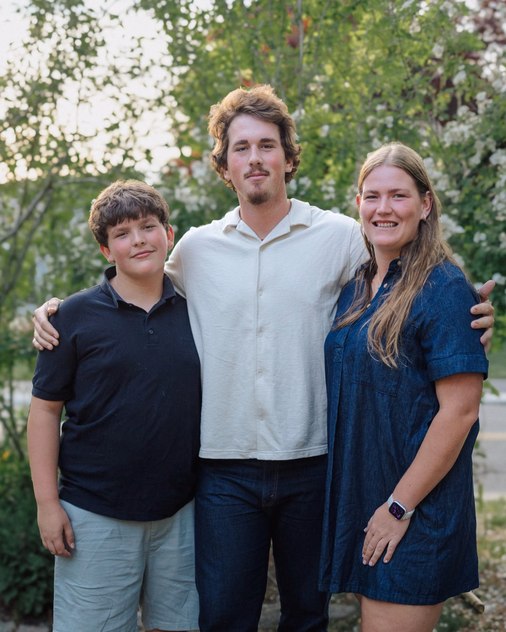 : Three people standing outdoors in front of green foliage, smiling at the camera. The person in the middle has an arm around the other two.