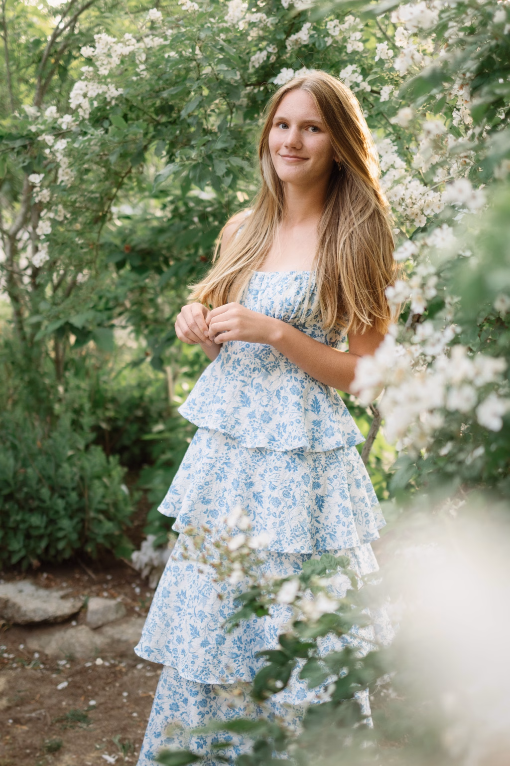 : Young woman with long blonde hair wearing a blue and white floral dress, standing outdoors surrounded by blooming white flowers and green foliage.