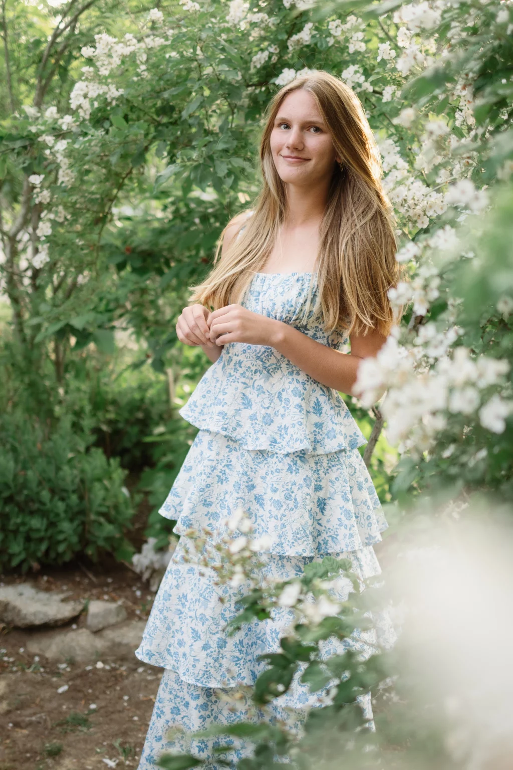: Young woman with long blonde hair wearing a blue and white floral dress, standing outdoors surrounded by blooming white flowers and green foliage.