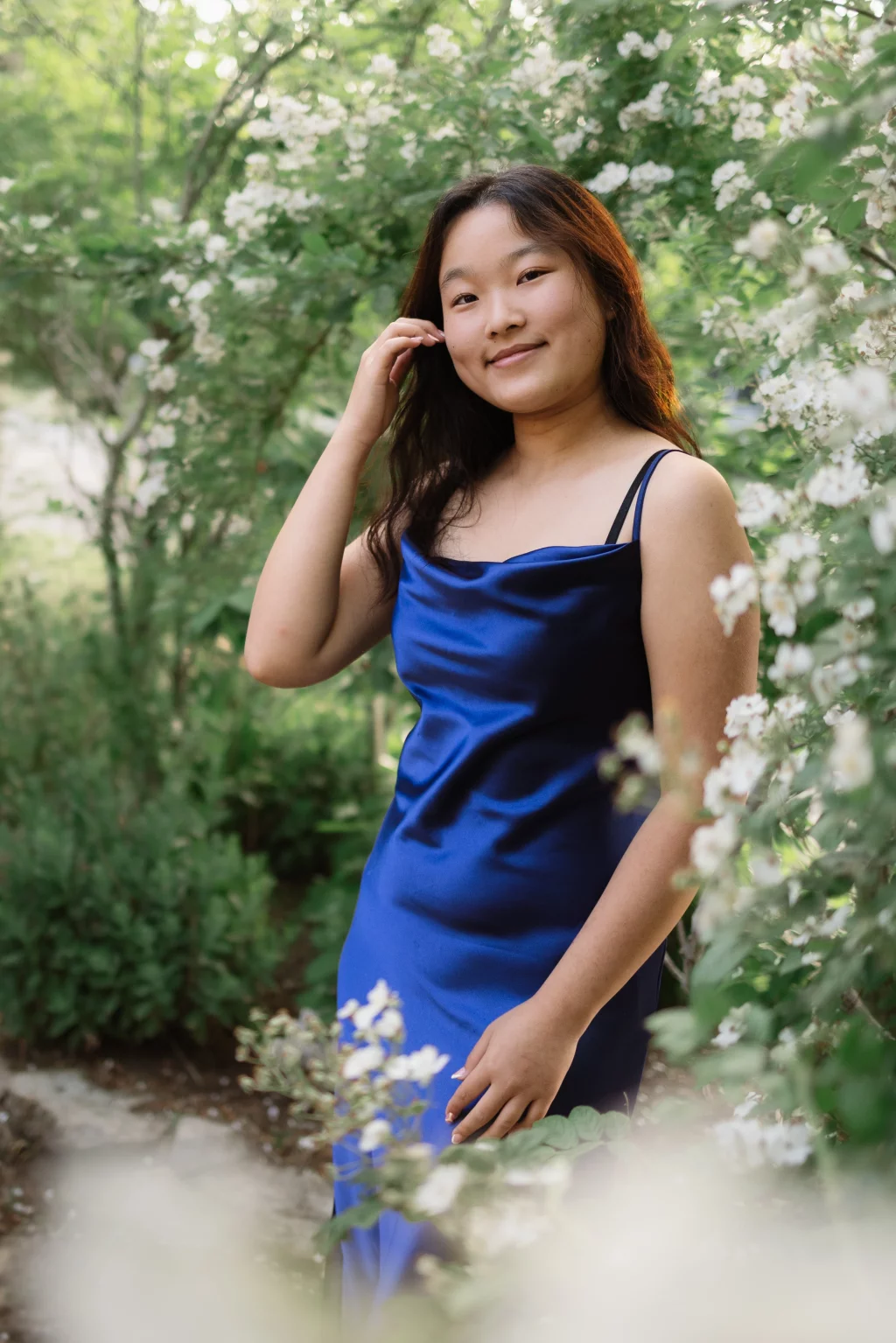 : A young woman in a blue satin dress stands among green foliage and white flowering bushes, smiling gently at the camera.
