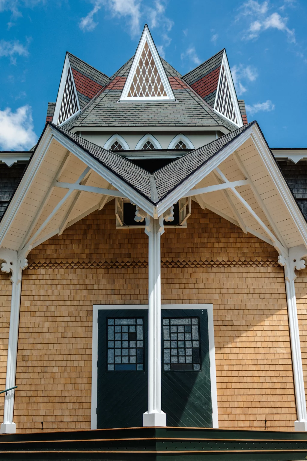 : Close-up of a house with tan wooden shingles, a dark green door with decorative glass panes, white trim, and a steep, pointed roof featuring triangular dormer windows with diamond-patterned glass under a blue sky.