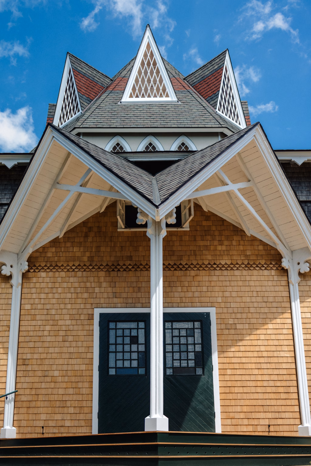 : Close-up of a house with tan wooden shingles, a dark green door with decorative glass panes, white trim, and a steep, pointed roof featuring triangular dormer windows with diamond-patterned glass under a blue sky.