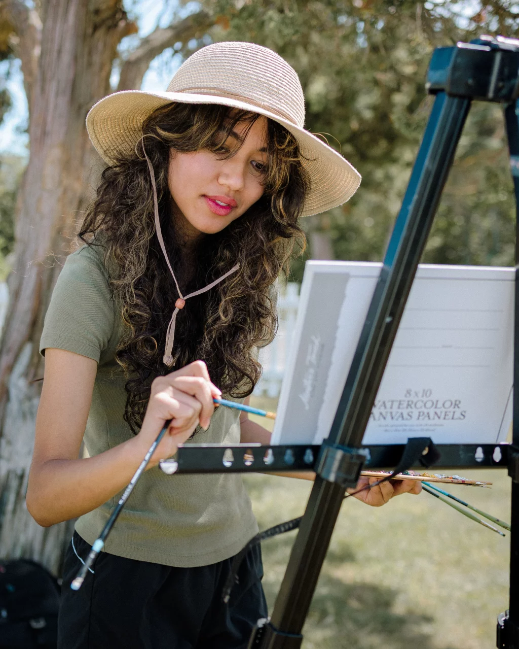 : Young woman wearing a wide-brimmed hat painting outdoors at an easel, holding brushes and a palette, with trees in the background.