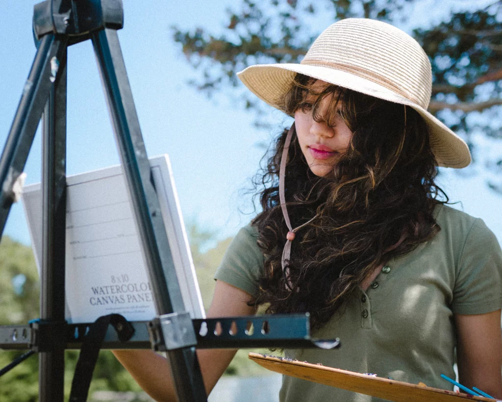 : Young woman wearing a wide-brimmed hat and green shirt painting outdoors at an easel, holding a palette.