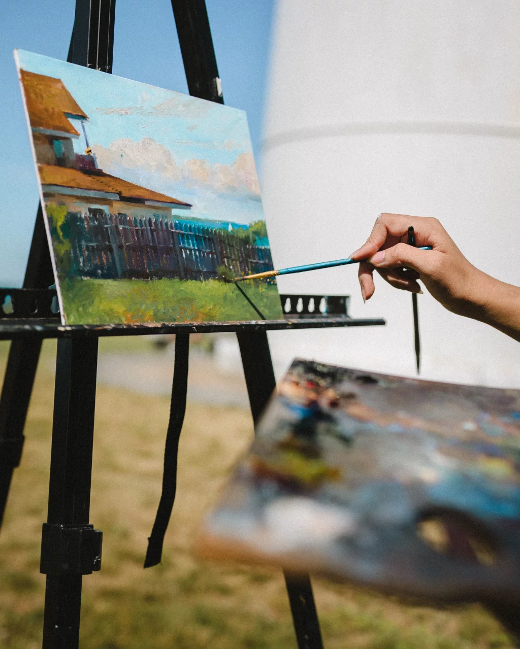 : A hand paints a landscape scene of a house, fence, and grass on a canvas set on an easel outdoors, with a palette visible in the foreground.