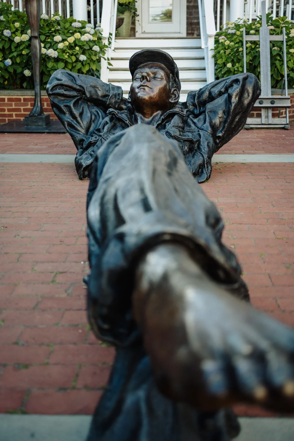 : Bronze statue of a person reclining with hands behind their head and one leg outstretched, wearing casual clothing and a cap, situated on a brick walkway in front of a building with white stairs and hydrangea bushes.