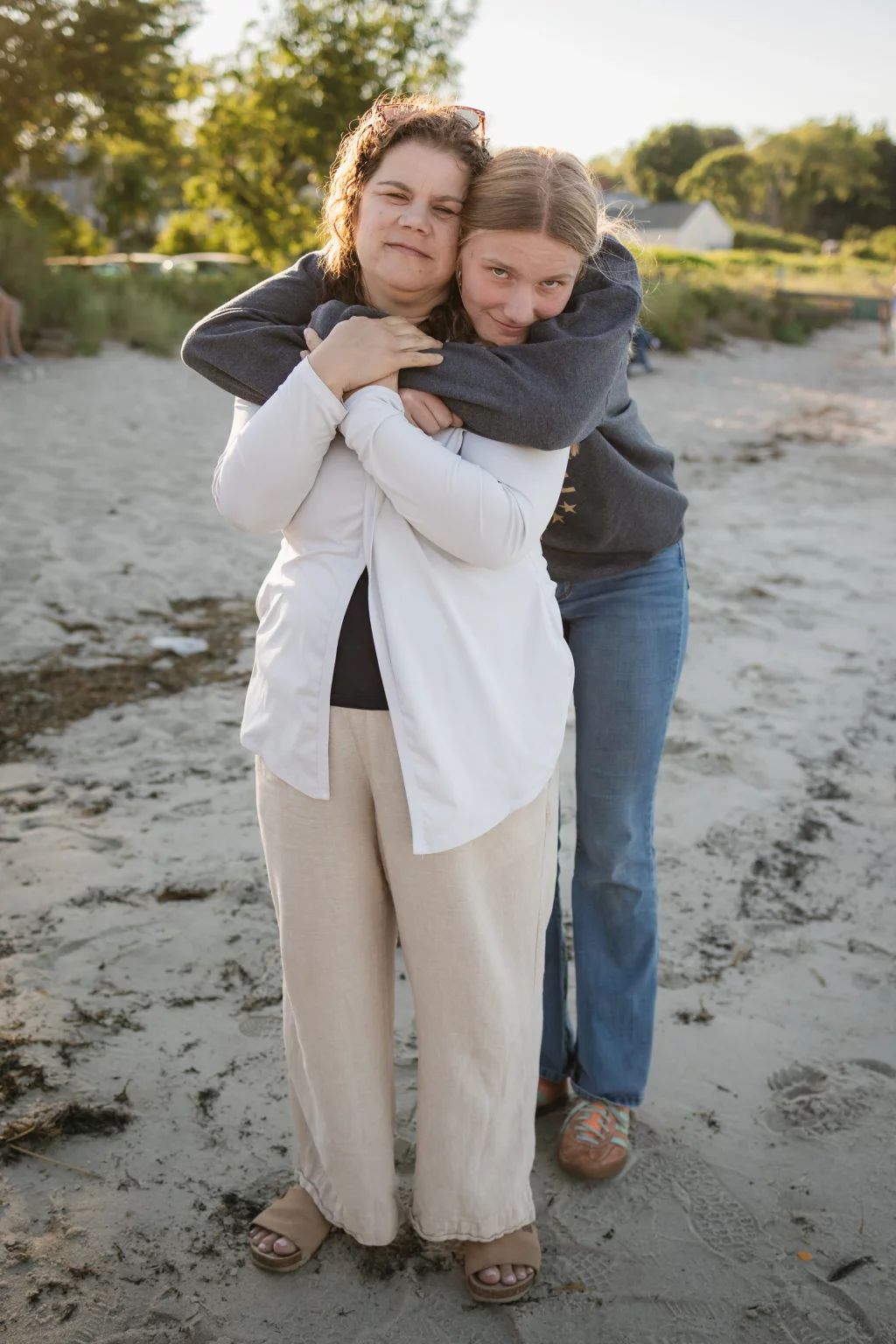 : Two people standing on a sandy beach; the person in front wears light pants and a white cardigan, while the person behind, dressed in jeans and a dark sweatshirt, hugs them from behind with arms around their shoulders. Both look at the camera with relaxed expressions. Trees and greenery appear in the background.