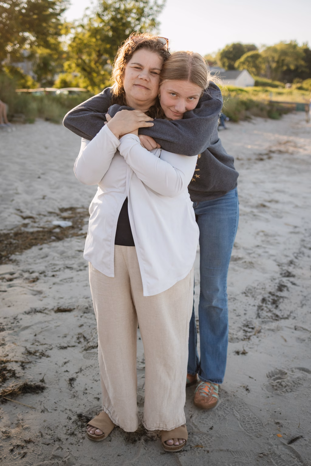 : Two people standing on a sandy beach; the person in front wears light pants and a white cardigan, while the person behind, dressed in jeans and a dark sweatshirt, hugs them from behind with arms around their shoulders. Both look at the camera with relaxed expressions. Trees and greenery appear in the background.
