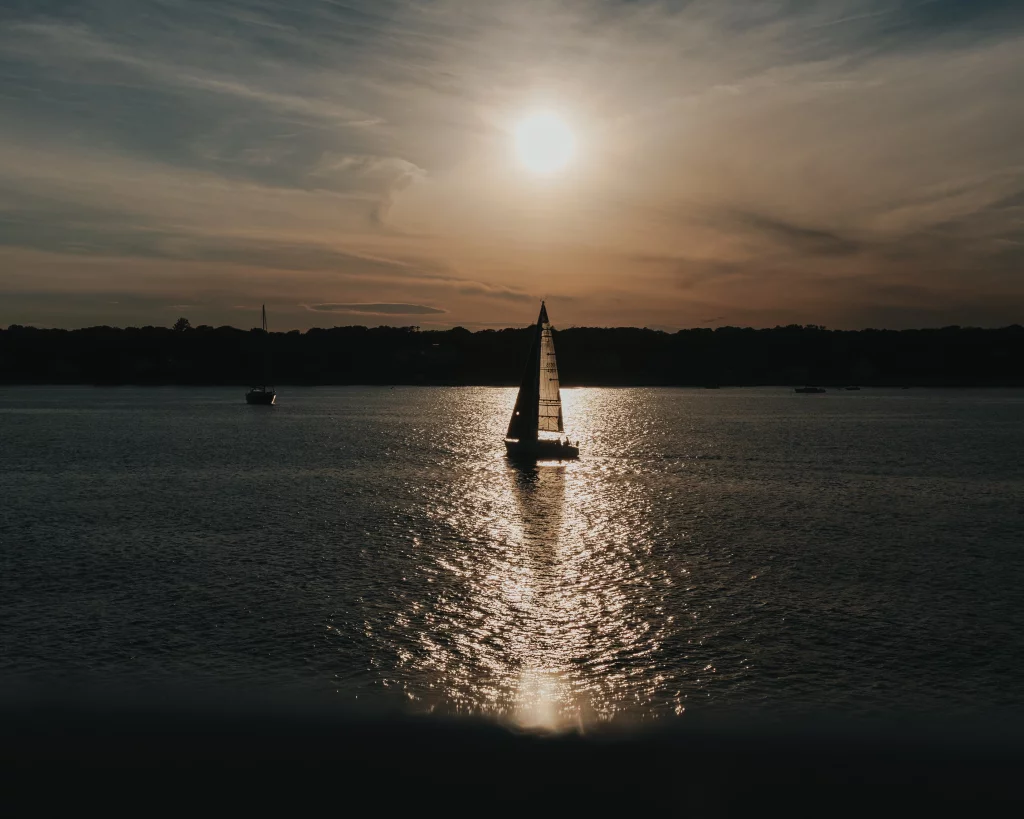 : Silhouette of a sailboat on calm water at sunset, with the sun low in the sky and its reflection shimmering across the surface.
