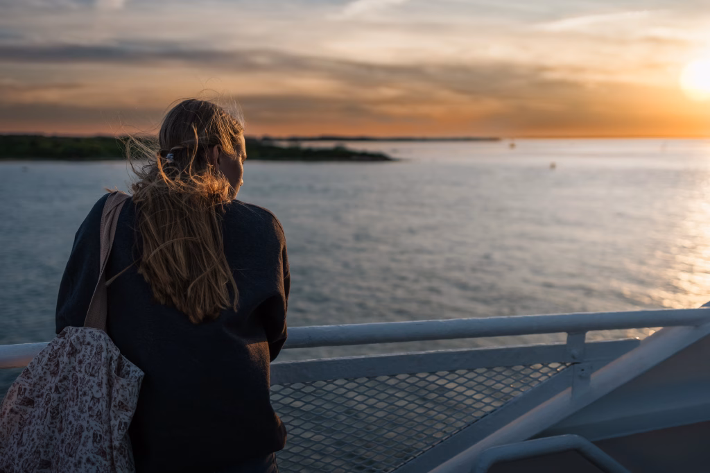 : Person with long hair standing on a ferry, looking out over the water at sunset with a patterned tote bag over their shoulder.