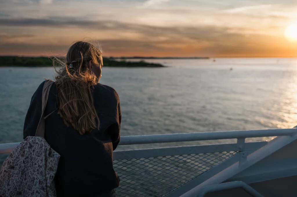 : Person with long hair standing on a ferry, looking out over the water at sunset with a patterned tote bag over their shoulder.
