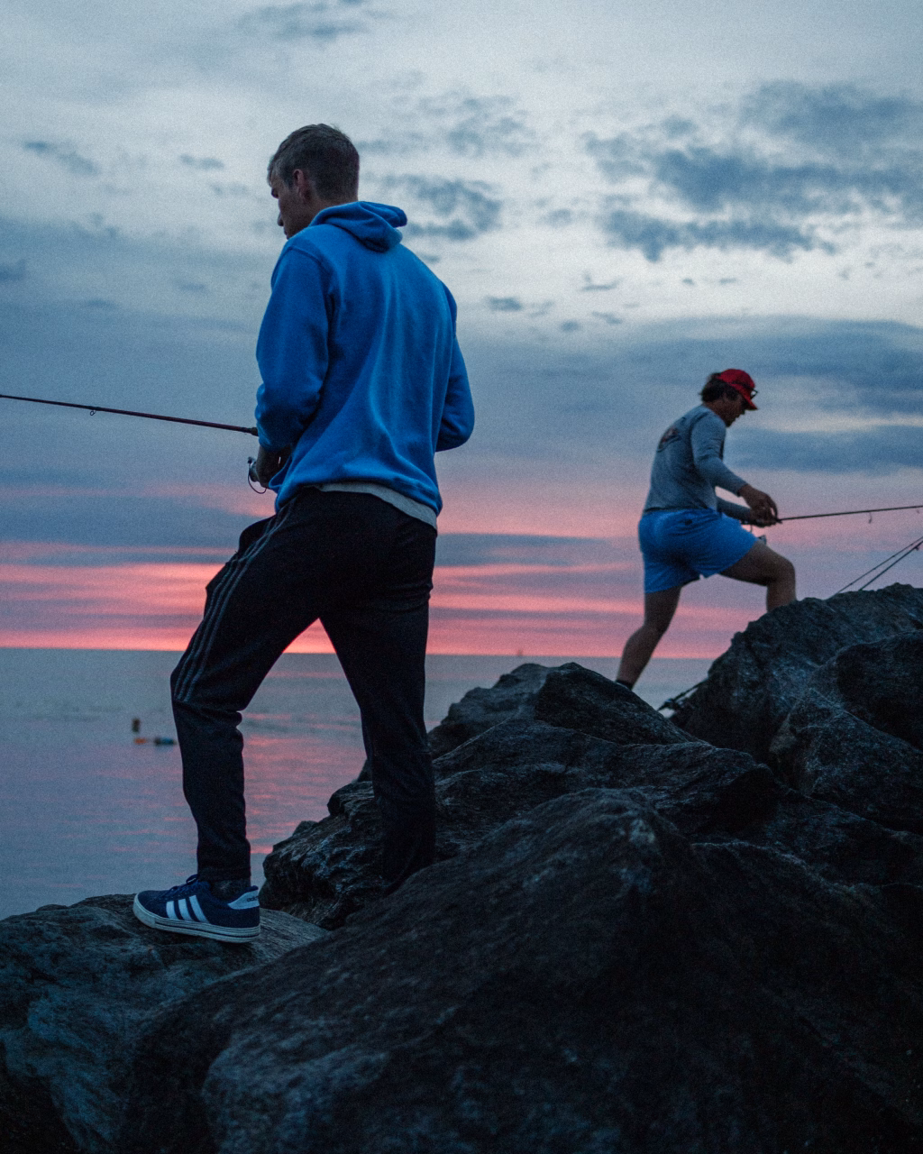 : Two people fishing on rocky shore at sunset, with a colorful sky and calm water in the background.