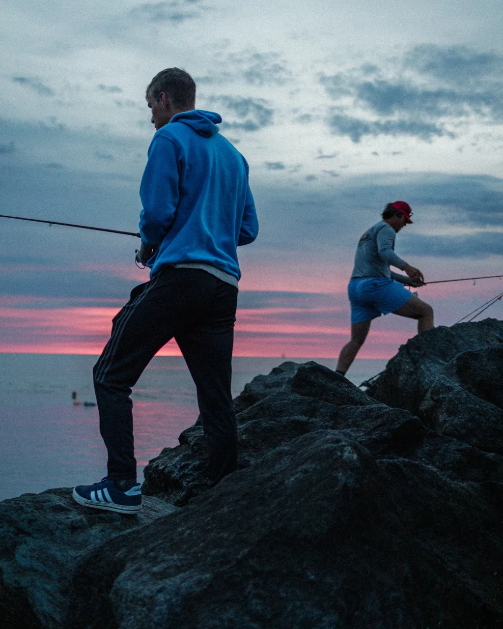 : Two people fishing on rocky shore at sunset, with a colorful sky and calm water in the background.
