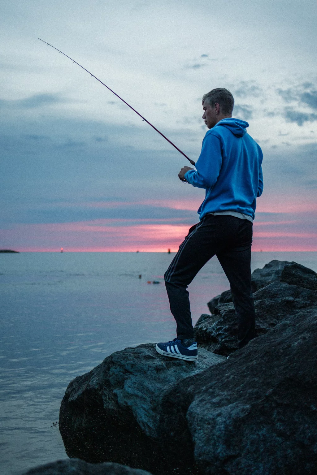 : Person in a blue hoodie fishing from rocks by the water at sunset, with a pink and purple sky in the background.