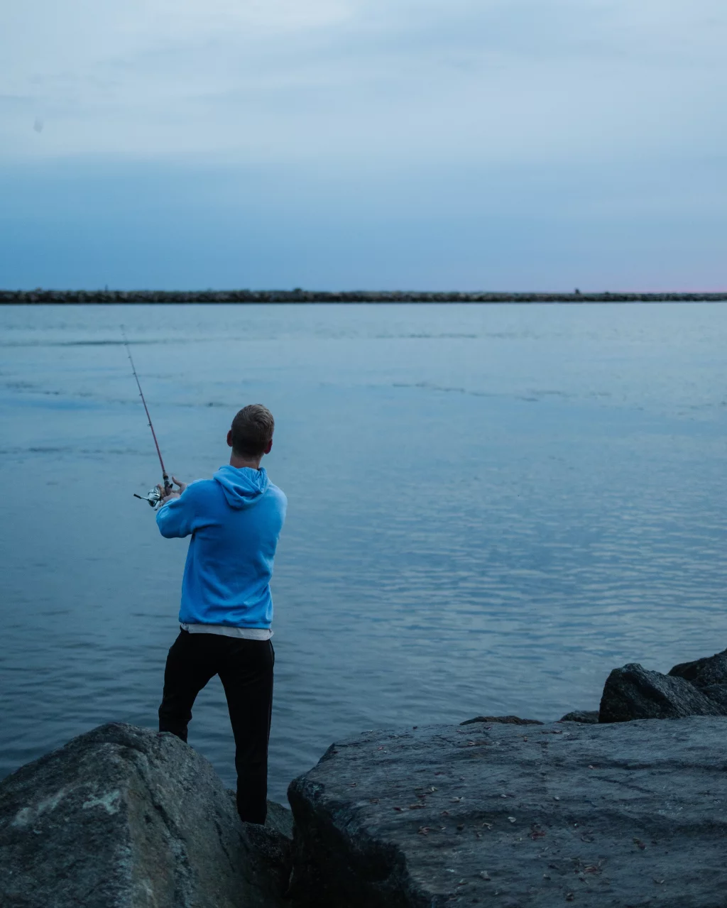 : Person in a blue hoodie fishing while standing on large rocks by a calm body of water under an overcast sky.