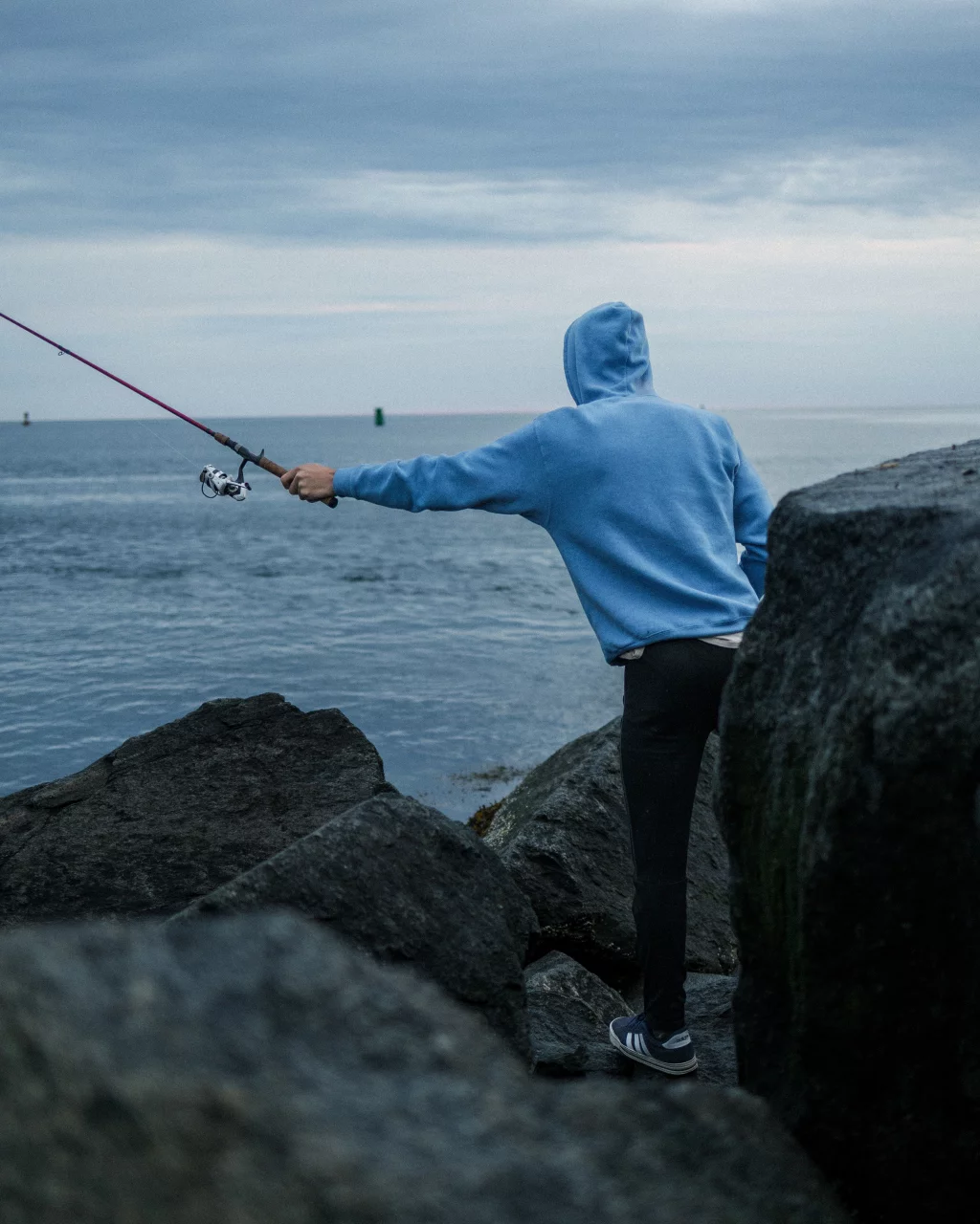 : Person in a blue hoodie fishing from large rocks by the water on a cloudy day.