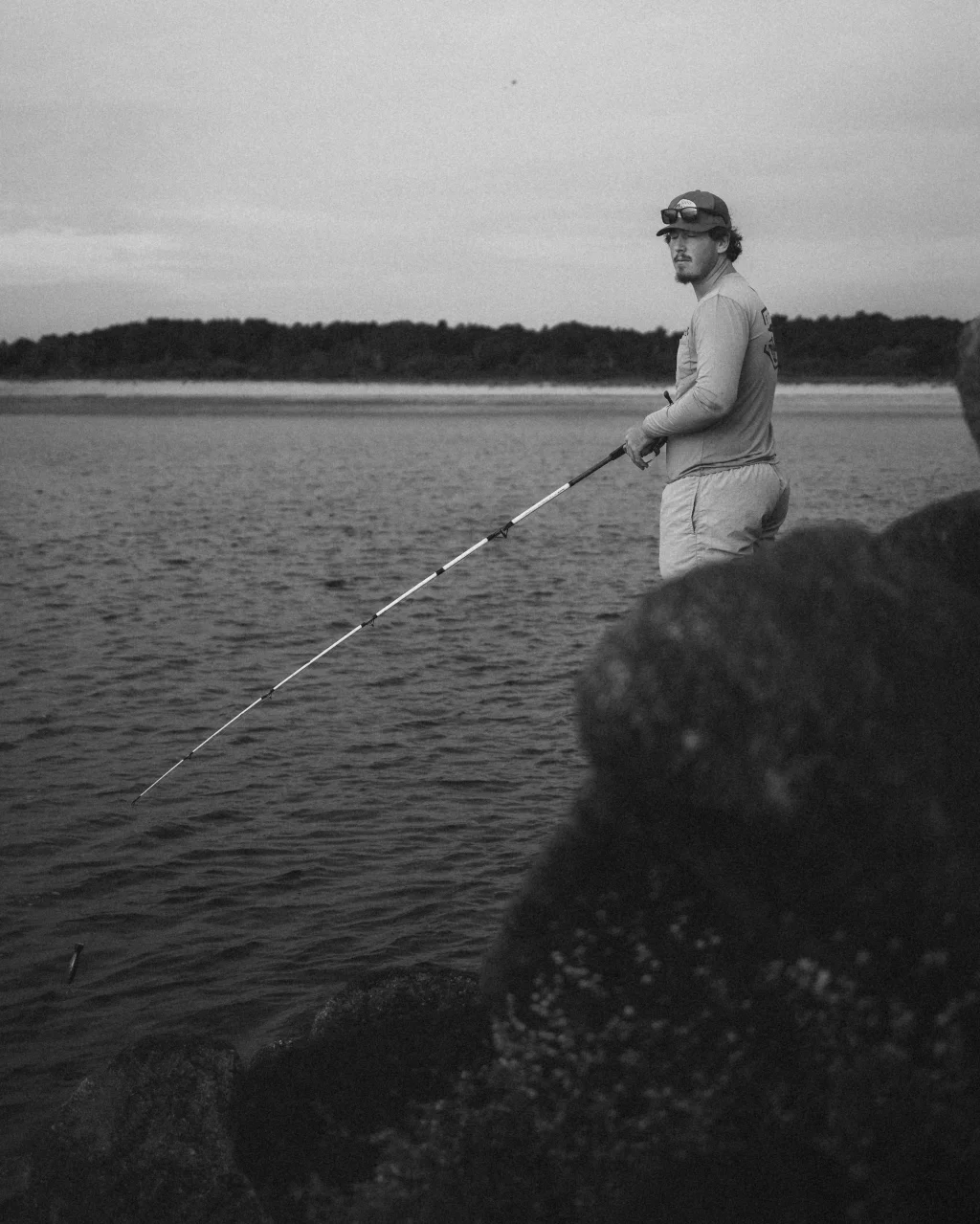 : Black and white photo of a person standing on rocks by the water, fishing with a rod, and looking toward the camera. Trees and shoreline are visible in the background.