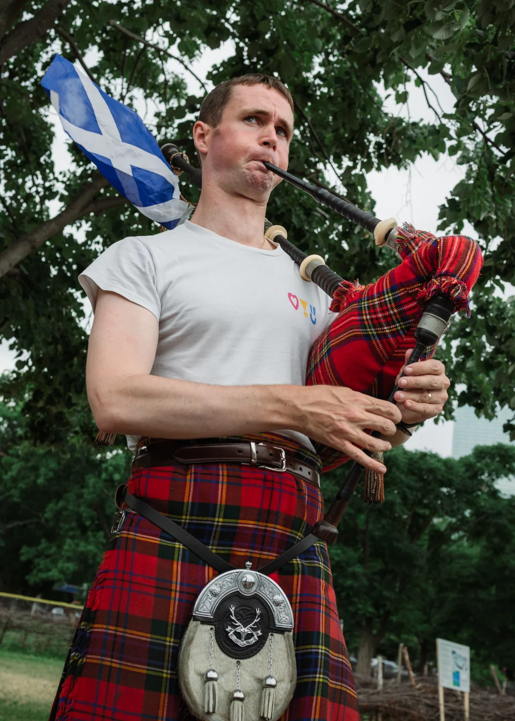 : A man wearing a red tartan kilt and white t-shirt plays the bagpipes outdoors, with a Scottish flag visible behind him and trees in the background.