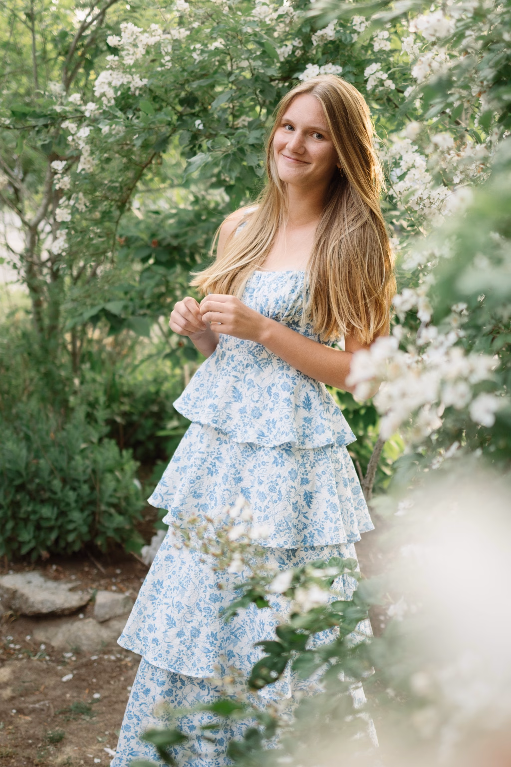 : Young woman with long blonde hair wearing a blue and white floral dress, standing outdoors among green bushes and white flowers, smiling softly.