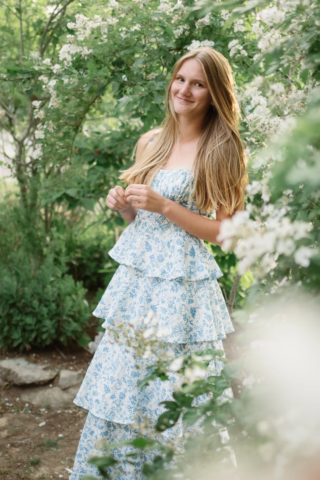 : Young woman with long blonde hair wearing a blue and white floral dress, standing outdoors among green bushes and white flowers, smiling softly.